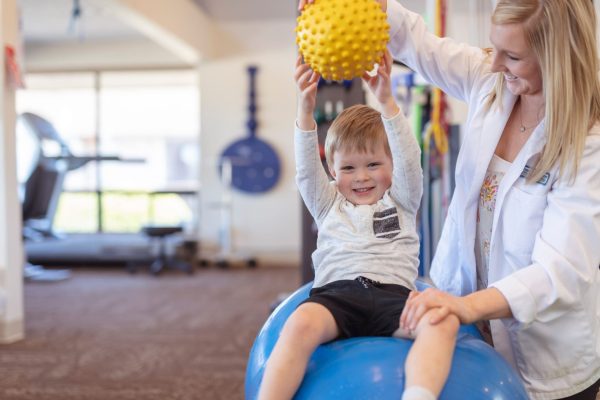 A blonde physical therapist works with a young boy in an open floor clinic.